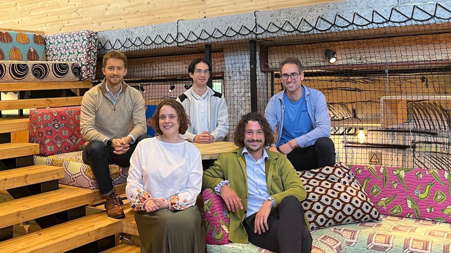 A group pf five people sitting beside a wooden staircase, on colourful cushions. Photo.