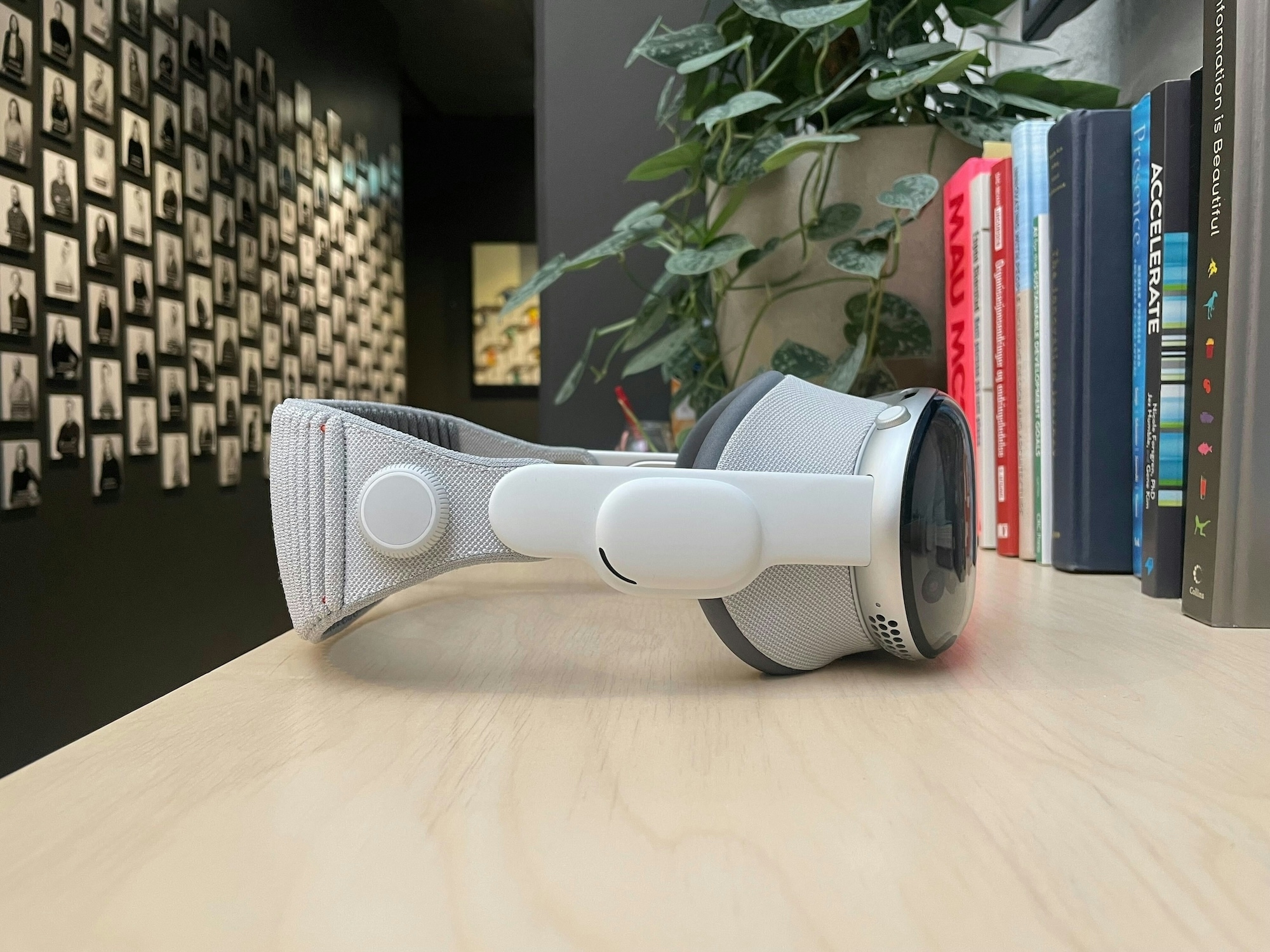 An Apple Vision Pro device placed on a wooden surface. A plant and dark wall with photos showing in the background. Photo and render.